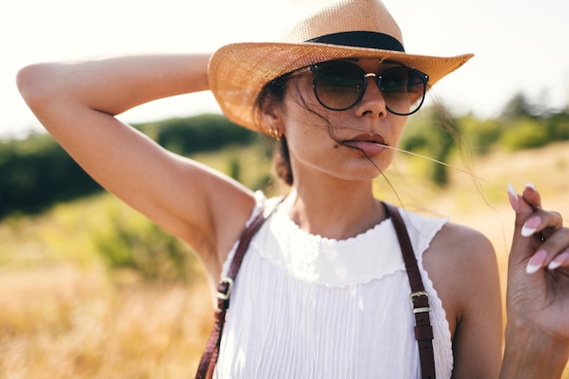 Espíritu de libertad Una atractiva chica boho con sombrero de blusa y gafas de sol de pie en el campo sobre el fondo de un cielo azul Vacaciones de verano viajando al estilo hippie moderno bohemio