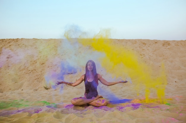 Espléndida mujer morena con cabello largo posando con una nube de pintura Holi, sentada en la arena