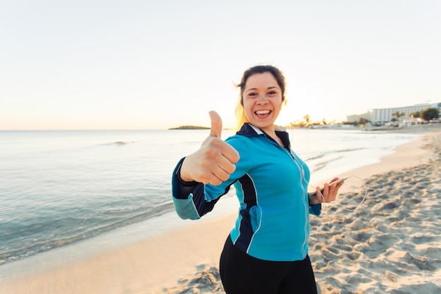 Esporte ao ar livre, dispositivo de fitness e conceito de pessoas - sorrindo fitness feminino mostra os polegares e segurando o smartphone com fones de ouvido.