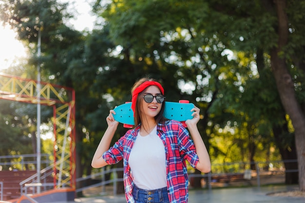 Esporte da juventude. mulher jovem hippie vestindo uma camisa xadrez vermelha e shorts jeans segurando um skate em um parque de skate. horário de verão