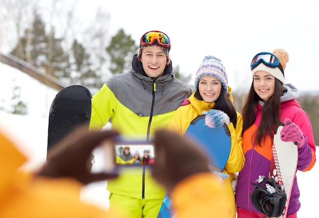 esporte de inverno, tecnologia, lazer, amizade e conceito de pessoas - amigos felizes com pranchas de snowboard e smartphone tirando fotos ao ar livre