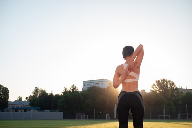 Esporte e bem estar. Garota fitness de tênis branco, fazendo exercícios de alongamento. Moda mulher desportiva com forte treinamento de corpo musculoso. Feminino apto alongando-se em estádio ao ar livre