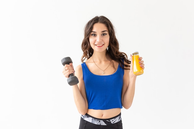 Esporte, estilo de vida saudável, conceito de pessoas - close-up de uma jovem com um haltere na mão e uma garrafa de suco na outra mão sobre fundo branco