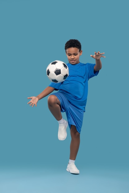 Esporte favorito. Menino persistente de pele escura usando shorts e camiseta jogando bola de futebol com o joelho para cima sobre fundo azul