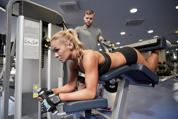 esporte, fitness, musculação, trabalho em equipe e conceito de pessoas - jovem e personal trainer flexionando os músculos na máquina de flexão de pernas no ginásio