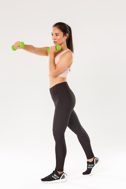 Esporte, ginástica e conceito de corpo saudável. comprimento total de athelte feminino focado, menina morena asiática, fazendo exercícios de fitness, treino com halteres, treinamento de ginástica produtivo sobre fundo branco.