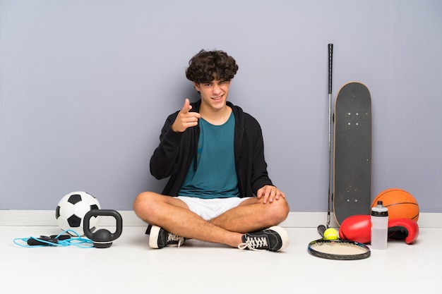 Esporte jovem homem sentado no chão em torno de muitos elementos do esporte aponta o dedo para você