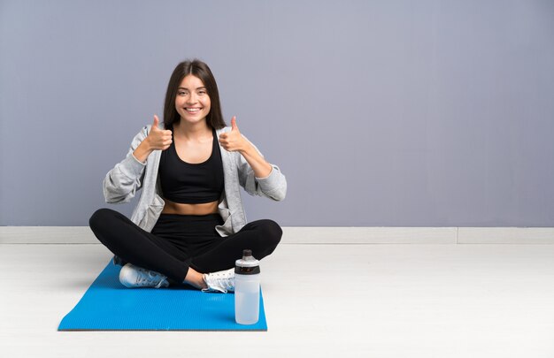 Esporte jovem mulher sentada no chão com tapete dando um polegar para cima gesto