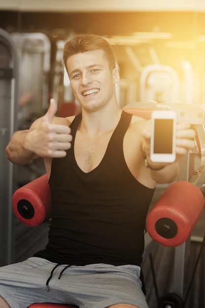 esporte, musculação, estilo de vida, tecnologia e conceito de pessoas - jovem sorridente mostrando smartphone e polegares para cima gesto no ginásio