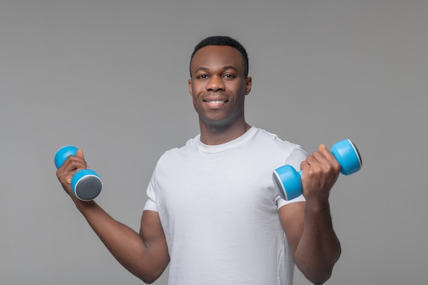 Esportes, halteres. Jovem adulto esportivo afro-americano sorridente com camiseta branca em pé com halteres nas mãos