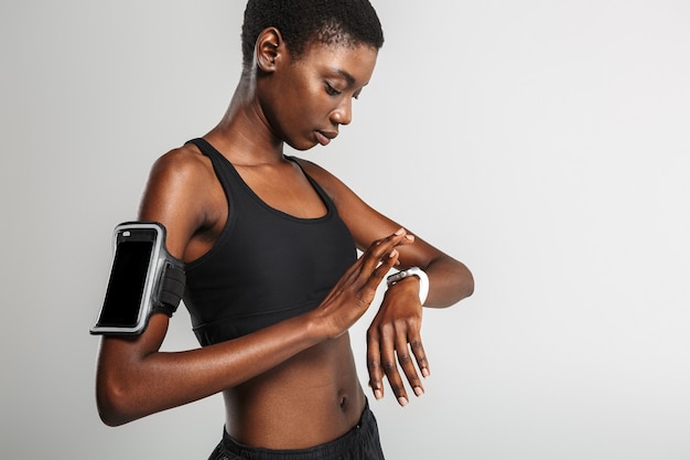 Esportista afro-americana usando celular e smartwatch enquanto treina isolada na parede branca