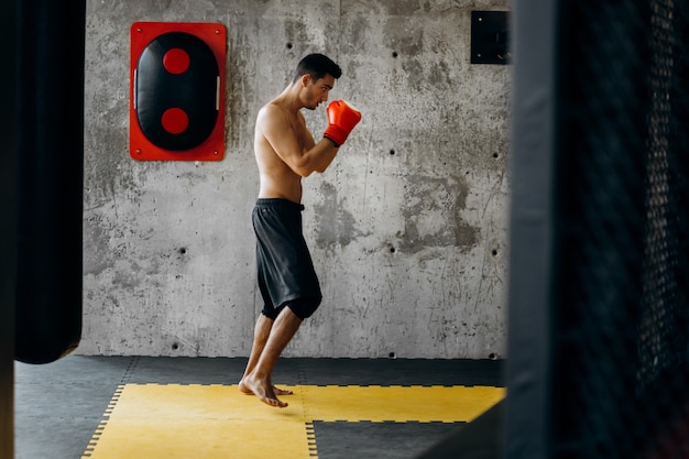 Esportista com o torso nu e com as luvas de boxe vermelhas ataca o equipamento de boxe em uma parede de concreto do ginásio.