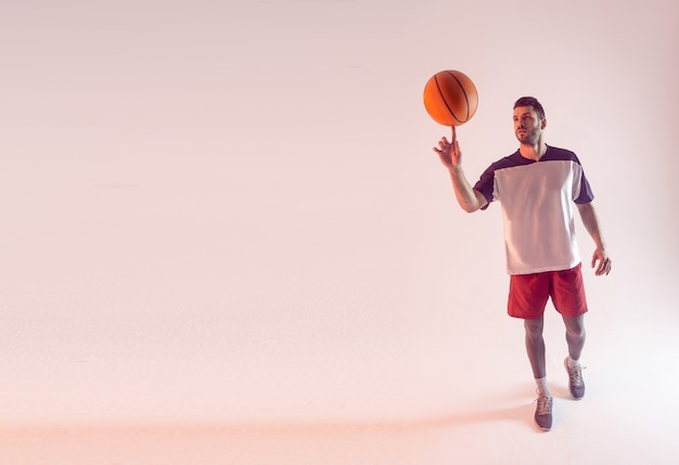 Esportista concentrado girando a bola de basquete no dedo. Jovem jogador de basquete europeu bonito barbudo usar roupas esportivas. Isolado em fundo bege. Sessão de estúdio. Copie o espaço