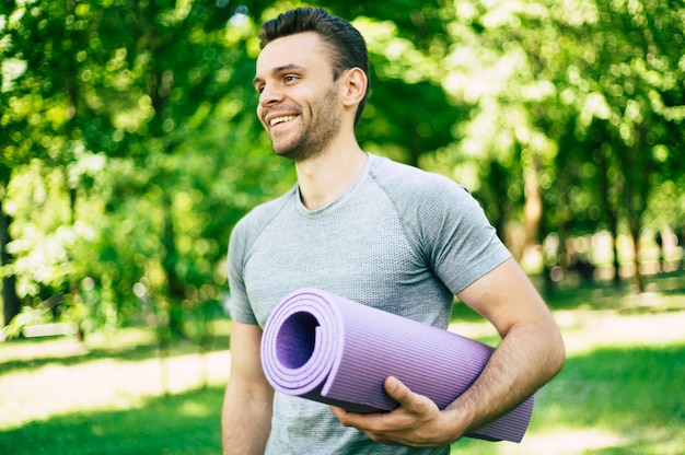 Esportista desportivo, magro e forte bonito ou instrutor de ioga com tapete de ioga na mão no fundo do parque da cidade