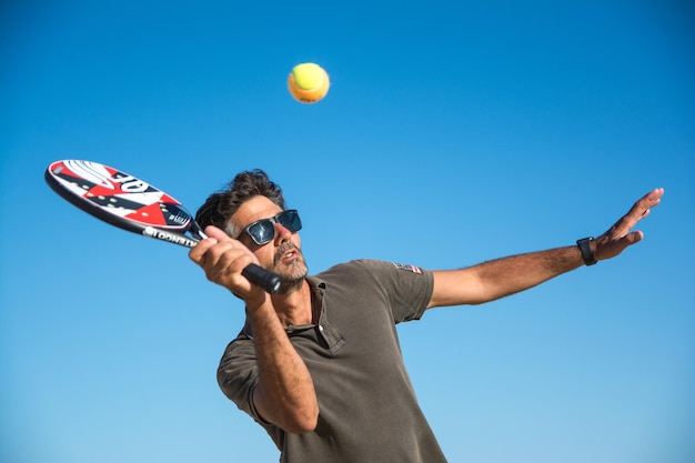 Esportista jogando tênis na praia com fundo de céu azul. Jogador de Paddle