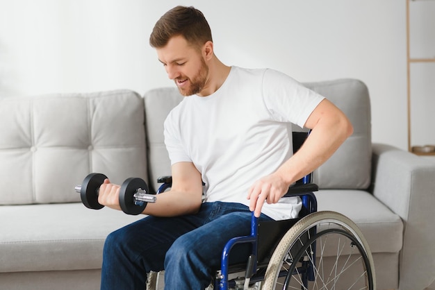 Esportista sentado em cadeira de rodas e braços estendidos com halteres durante o exercício de reabilitação no centro médico moderno Homem sentado na cadeira de rodas em casa