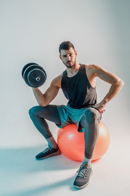 Esportista, treinamento de bíceps com halteres na bola de fitness. jovem confiante barbudo homem europeu usa uniforme de esportes e olhando para a câmera. isolado em fundo turquesa. sessão de estúdio