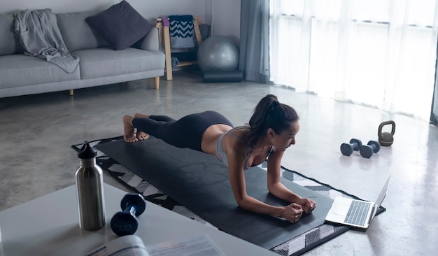 Esportiva jovem asiática fazendo prancha na frente do laptop sala de estar interior vista lateral espaço de cópia Jovem saudável em roupas esportivas se exercitando em casa assistindo aula de fitness on-line