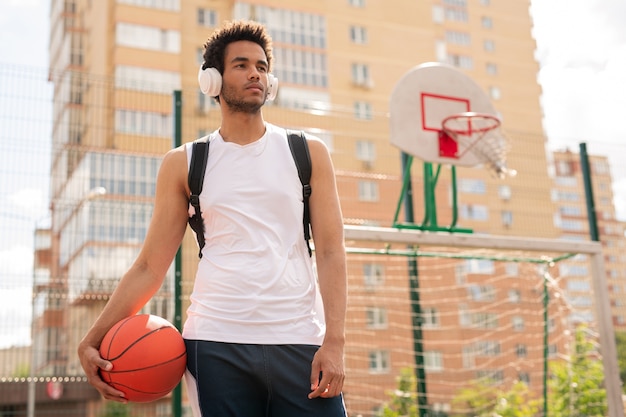 Esportivo com bola para jogar basquete, ouvindo música em fones de ouvido no parquinho do campo