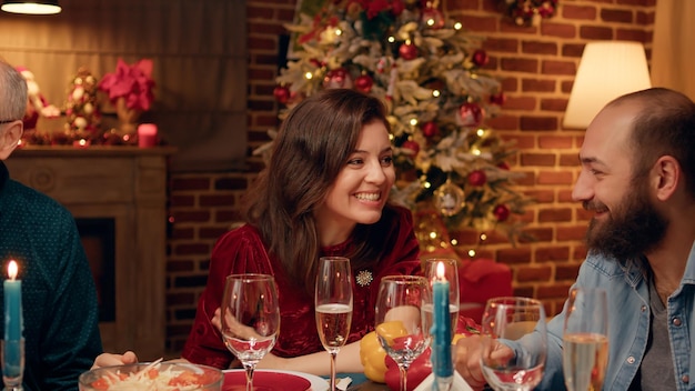 Esposa festiva hablando con su marido mientras celebra la Navidad en la mesa familiar de la cena. Linda mujer sonriendo sinceramente a la cámara mientras disfruta de las vacaciones de invierno con familiares cercanos.