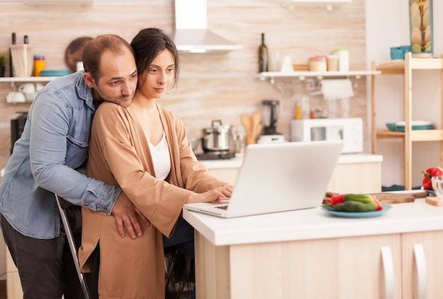 Esposa lendo no laptop na cozinha enquanto o marido a abraça. Casal feliz, amoroso, alegre, romântico, apaixonado, em casa, usando a moderna tecnologia de internet wi-fi sem fio