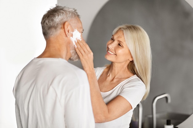 Esposa madura amorosa a aplicar espuma de barbear no rosto do marido na casa de banho