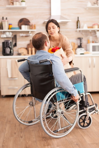 Esposa pegando sacola de supermercado do marido deficiente em cadeira de rodas depois de chegar do supermercado na cozinha. Homem deficiente paralisado e deficiente com deficiência motora que se integra após um acidente.