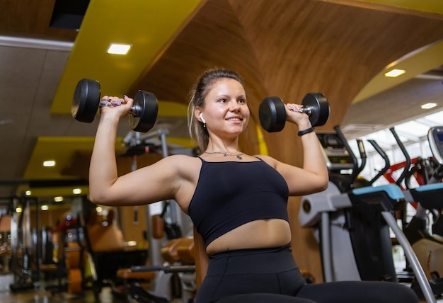 Esposa sorridente treina com halteres sentados em um banco na academia Treinamento muscular do ombro