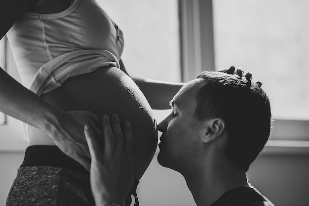 Un esposo besa el estómago de su esposa embarazada cerca de la ventana de la casa Expectativa de embarazo familiar feliz Foto en blanco y negro
