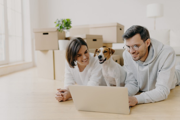 El esposo y la esposa alegres ven la película en línea en la computadora portátil, descansan en el piso, se relajan y hablan, sus animales domésticos posan entre ellos, se mudan a un nuevo hogar, posan en una espaciosa sala de estar con cajas desempaquetadas