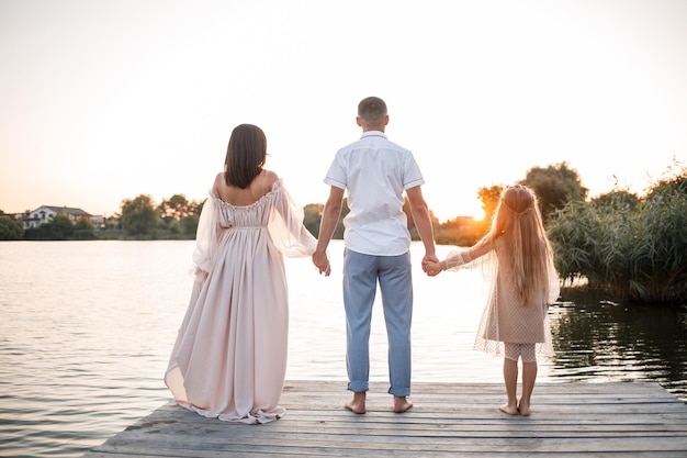 Un esposo con una esposa embarazada y una hija descansan en la orilla del río parados en el muelle en un clima soleado de verano al atardecer pareja enamorada hombre y mujer abrazándose tomados de la mano Una familia feliz