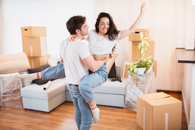 El esposo y la esposa felices se divierten y se trasladan al propio apartamento, concepto de reubicación. Joven pareja feliz baila en la sala de estar cerca de cajas de cartón entretiene en el día de la mudanza,