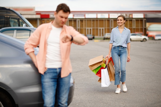 El esposo está esperando a su esposa en el estacionamiento del supermercado. Clientes felices con compras cerca del centro comercial, vehículos, pareja familiar en el mercado.