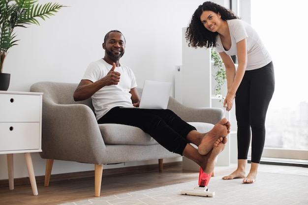 Esposo trabajando en una laptop mientras su esposa aspira el piso