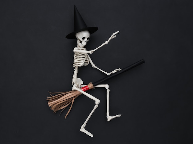 Esqueleto com chapéu de bruxa em uma vassoura. fundo preto. conceito minimalista de halloween. vista do topo. postura plana