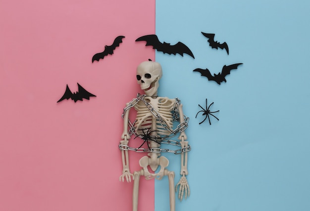 Esqueleto envolto em corrente de metal em pastel azul rosa com morcegos e aranhas. Decoração de Halloween, tema assustador