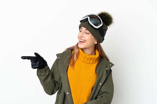 Esquiador mujer caucásica con gafas de snowboard aislado sobre fondo blanco apuntando con el dedo hacia el lado y presentando un producto