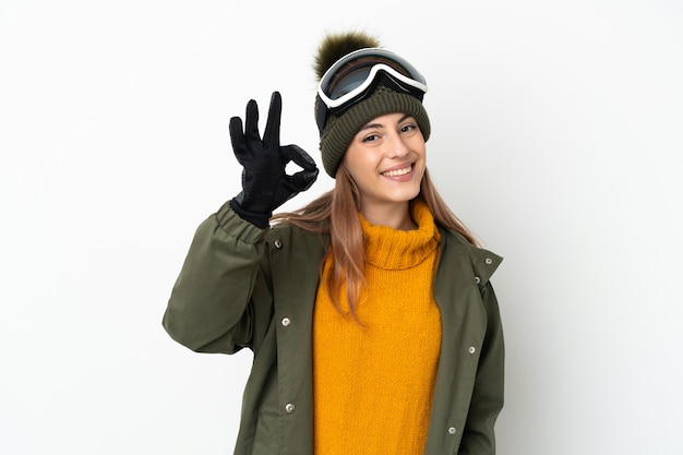 Esquiador mujer caucásica con gafas de snowboard en blanco mostrando signo ok con los dedos