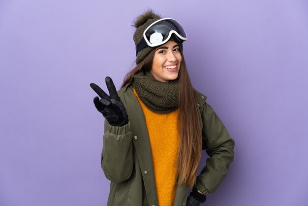 Esquiadora caucasiana com óculos de snowboard isolados em um fundo roxo sorrindo e mostrando sinal de vitória