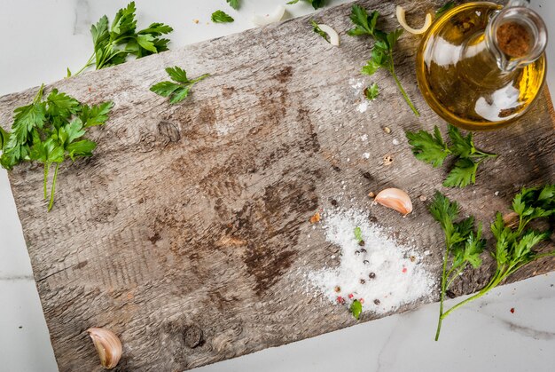 Essen kochen. Altes Schneidebrett auf einem weißen Marmorküchentisch. Olivenöl, ein Messer, Gewürze, Salz, Pfeffer, Knoblauch, Petersilie. Ansicht von oben