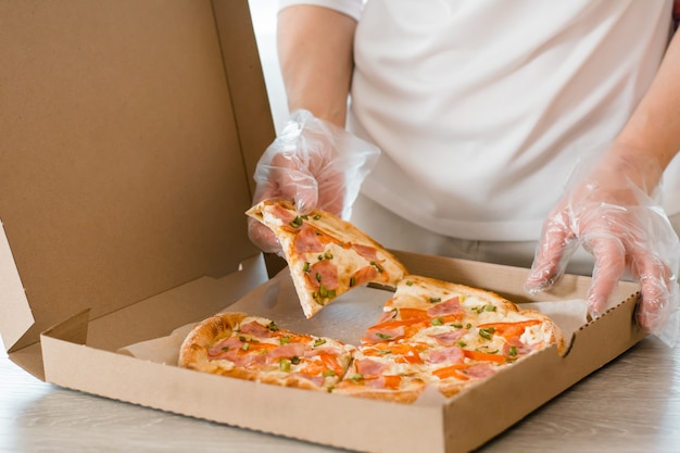 Essen zum Mitnehmen. Eine Frau in Einweghandschuhen nimmt ein Stück Pizza aus einem Karton auf dem Tisch in der Küche.