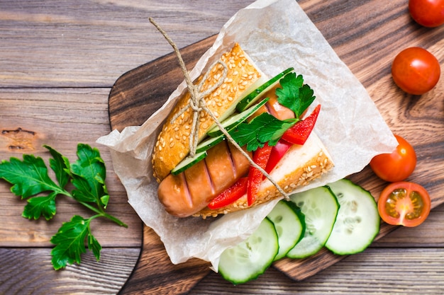 Essfertiger appetitanregender Hotdog gemacht von der gebratenen Wurst, von den Rollen und vom Frischgemüse, eingewickelt im Pergamentpapier auf einem Schneidebrett auf einem Holztisch