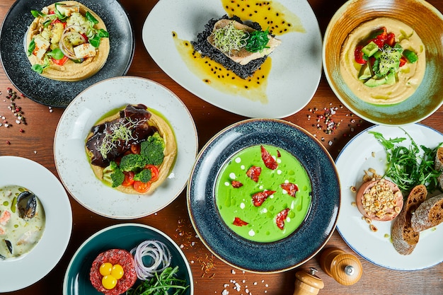 Esstisch mit ein paar Lebensmitteln. Grüne Sahnesuppe, Hummus, Dorado, Pastete, Bouillabaisse und Rindertartar für ein Familienessen. Draufsicht Essen flach lag.