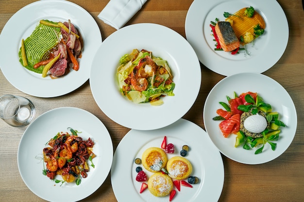 Esstisch mit einer großen Anzahl von Gerichten: Käsekuchen, Garnelensalat, Quinoa- und Lachssalat, Lammkarree, gebratene Meeresfrüchte, Lachssteak. Draufsicht Wohnung lag Essen