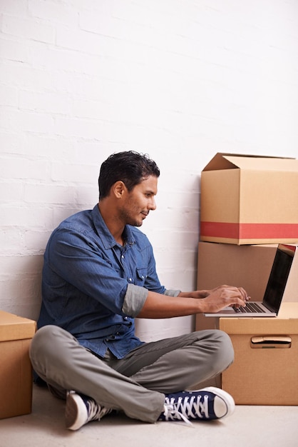 Se está mudando de casa Un apuesto joven usando su computadora portátil en el piso con cajas que lo rodean