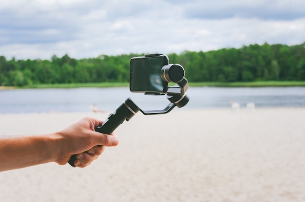 Estabilizador de câmera do smartphone na mão de um homem. no contexto de uma praia de areia e natureza com um lago.