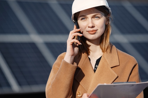 Estação de energia solar. jovem engenheira trabalha na fábrica. ela está falando ao telefone e fazendo negócios. mulher de capacete com papéis.