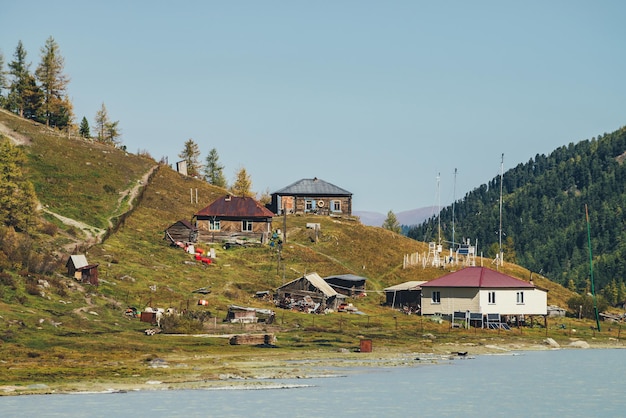 Estação meteorológica na colina perto do lago de montanha no vale entre as altas montanhas da floresta. Paisagem cênica de outono com aldeia de montanha na margem do lago e montanhas com floresta de coníferas. Três casas na colina