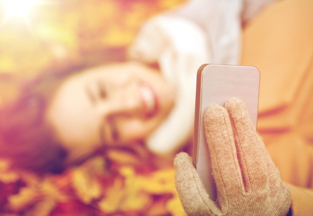Foto estação, tecnologia e conceito de pessoas - bela jovem deitada no chão e folhas de outono e tirando selfie com smartphone