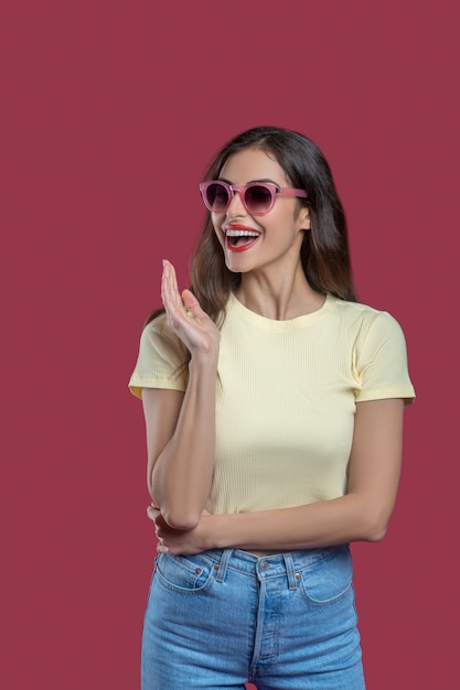 Estado de ánimo alegre. Feliz joven delgada en gafas de sol con la boca abierta en jeans y camiseta de pie sobre fondo rosa